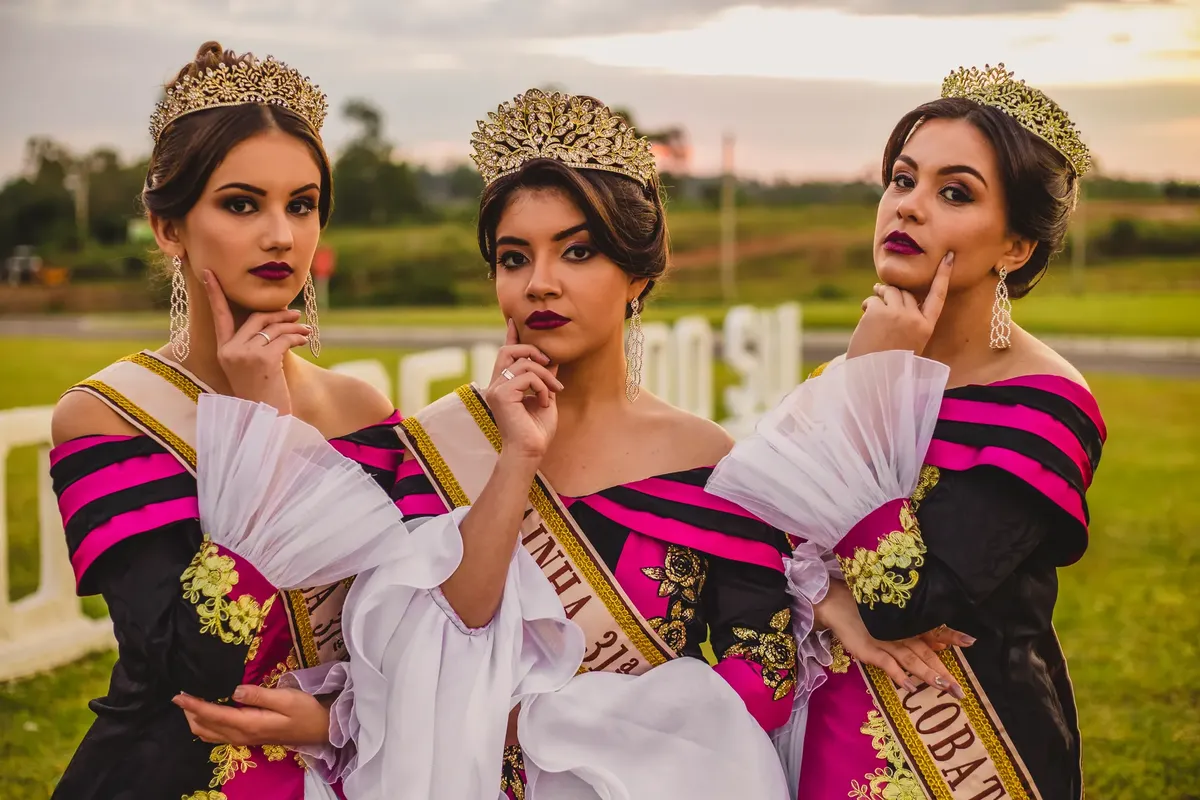 Three young women in elegant gowns and crowns posing outdoors for a pageant photoshoot.
