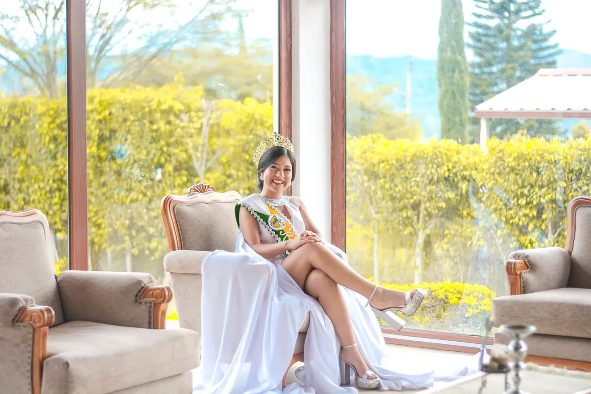 A beauty queen in a crown and sash sits elegantly in a luxurious room with large windows.