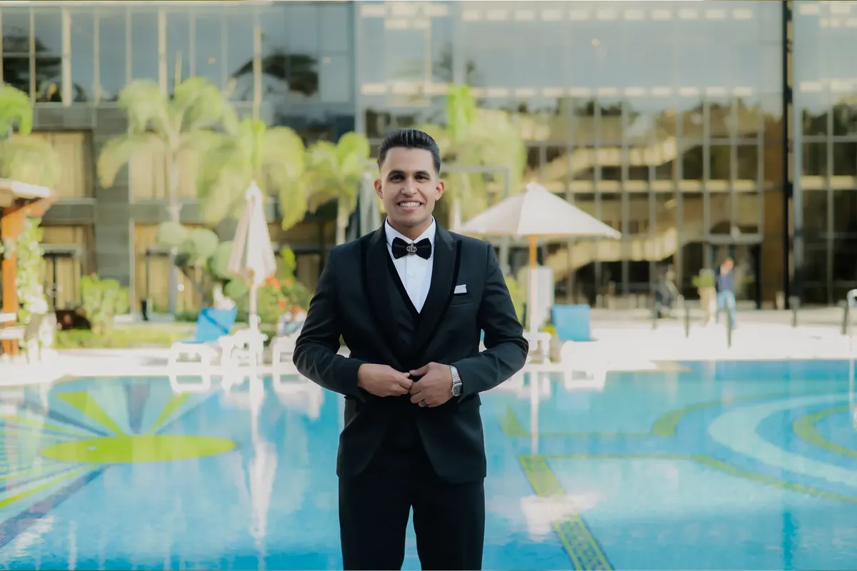 A confident man in a black tuxedo standing by a luxurious pool at a stylish outdoor venue.